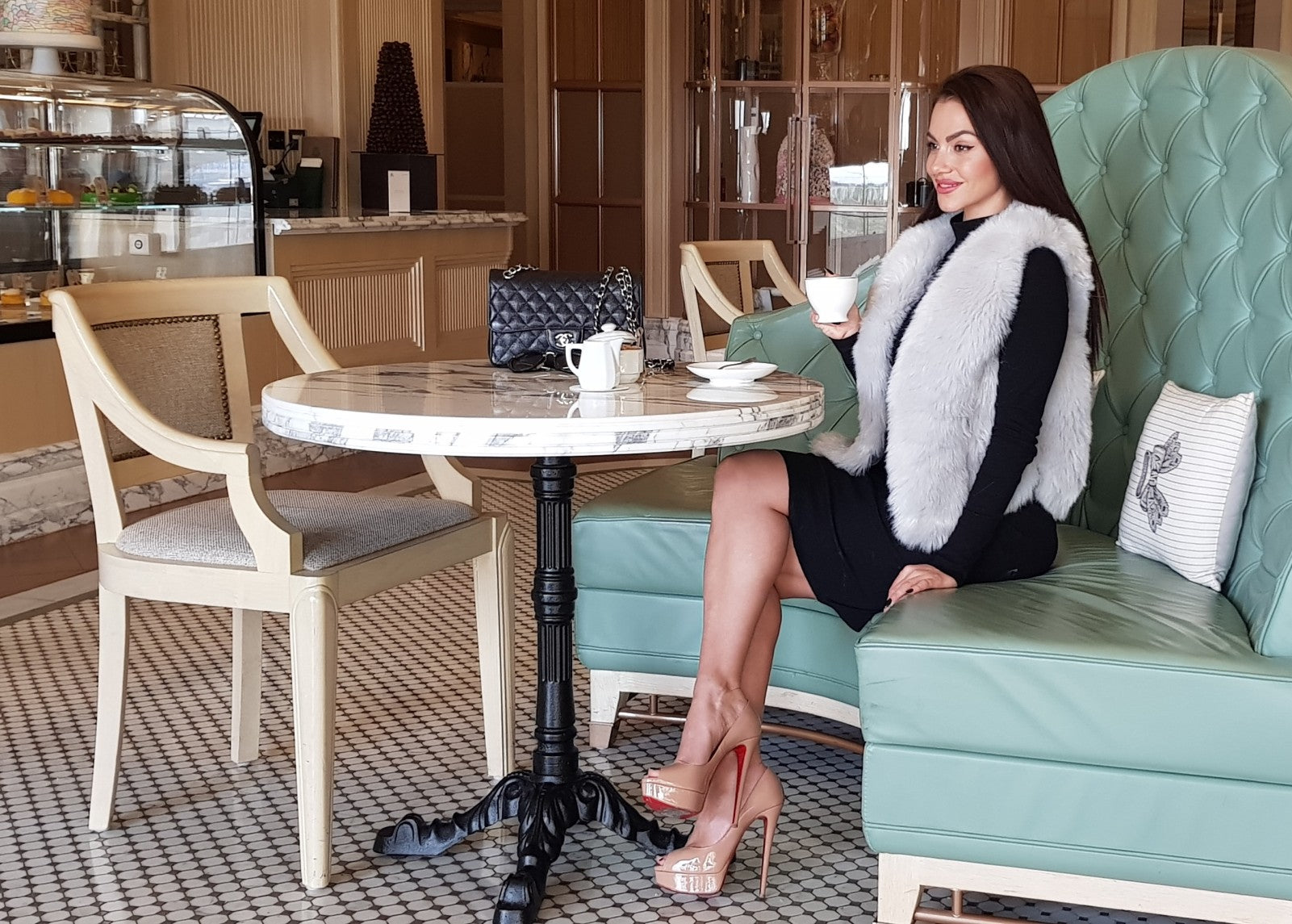 lovely young lady sitting at a marble table holding a cup of coffee and wearing a black dress and a short paris grey faux fur vest